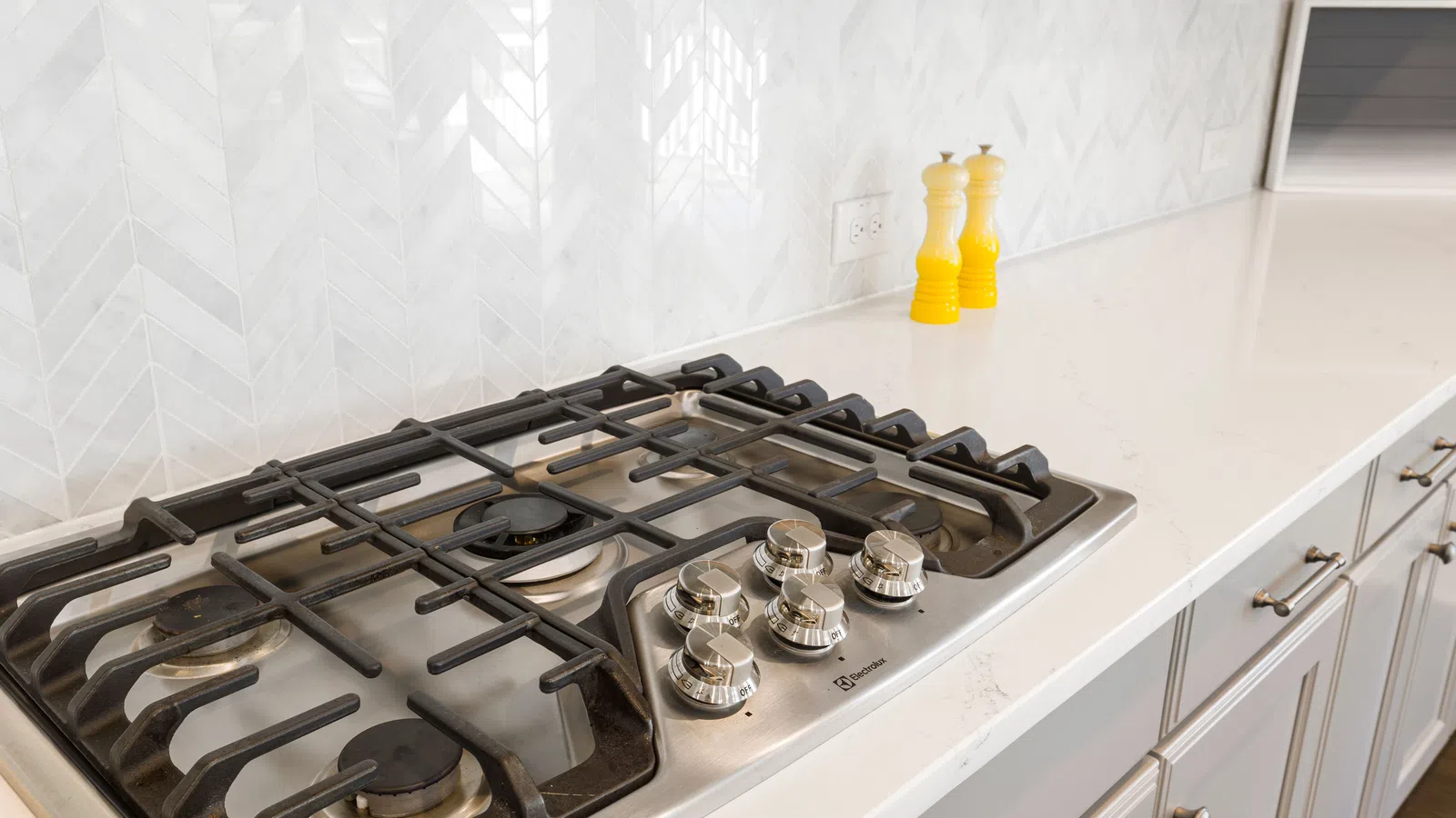 Modern stainless steel gas cooktop with four burners on a white kitchen countertop with yellow salt and pepper shakers.