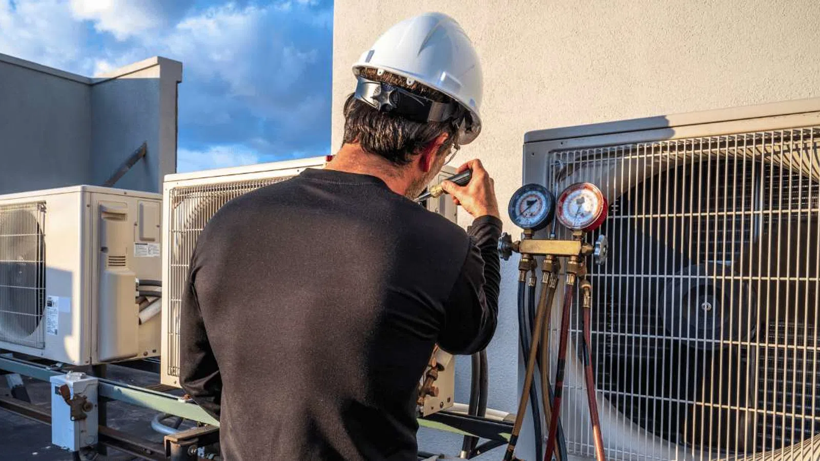 Technician in hard hat inspecting rooftop HVAC unit with pressure gauges under blue sky on sunny day
