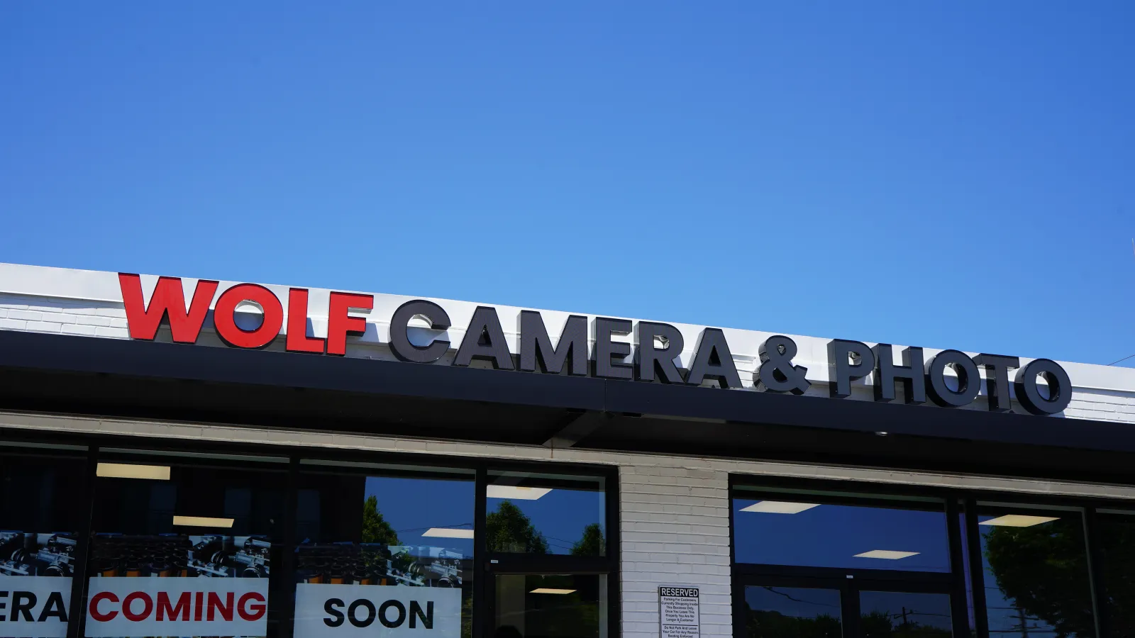 Storefront of Wolf Camera & Photo with a clear blue sky and Coming Soon signs in the windows.