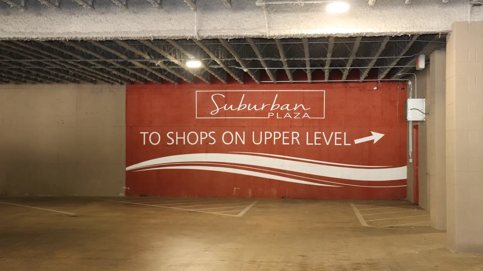 Red wall sign indicating shops are on the upper level in a parking garage with concrete ceiling.