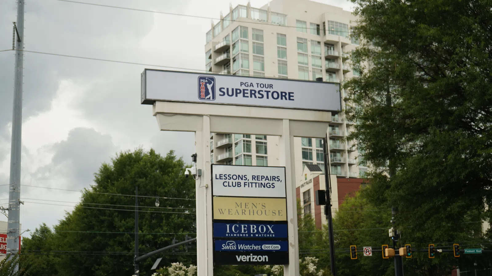 Outdoor multi-business sign for PGA Tour Superstore, Men's Wearhouse, Icebox, Verizon with city buildings and trees.