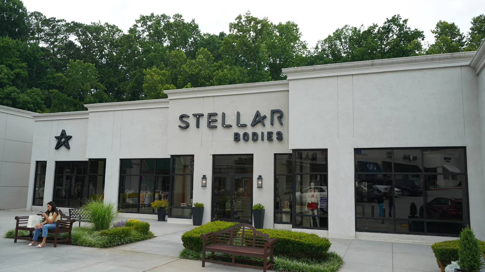 Modern white commercial building with Stellar Bodies signage, large windows, benches, greenery, and a person sitting outside.