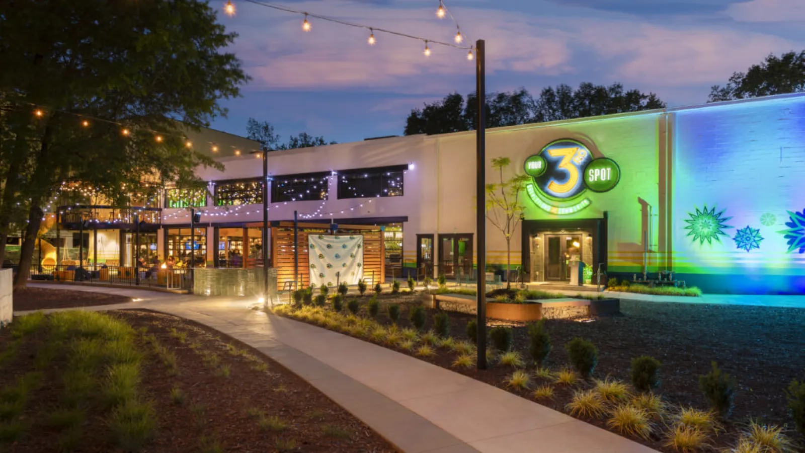 Evening view of a lit modern brewery with outdoor seating, string lights, and landscaped walkway at dusk.