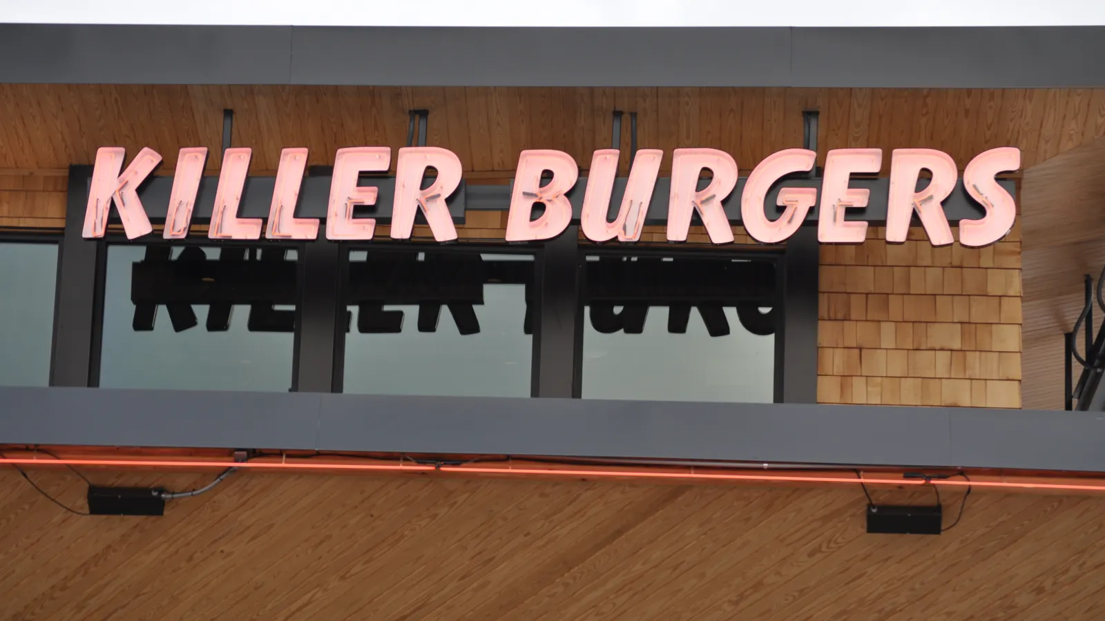 Bright pink neon sign reading Killer Burgers mounted on wooden exterior above windows.