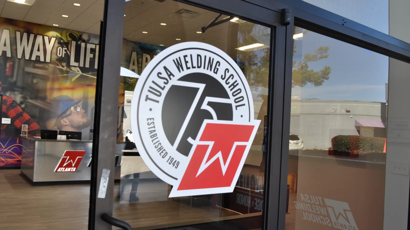 Entrance glass door with Tulsa Welding School logo and interior showing workspace and wall art