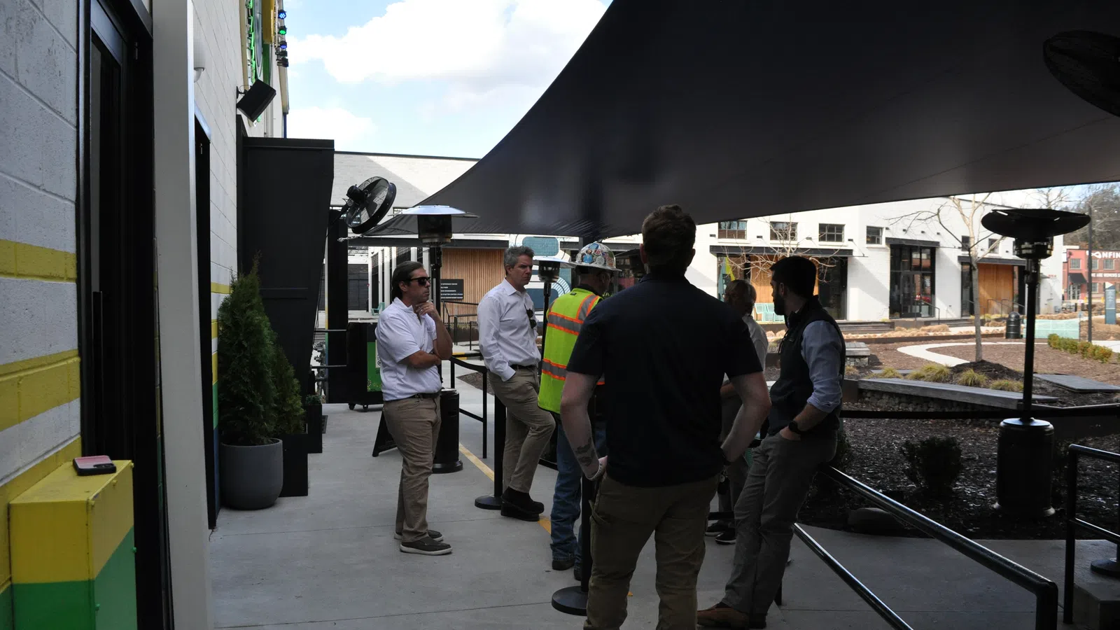 Group of people, including one in a safety vest, talking under a large black canopy outside modern buildings on a sunny day