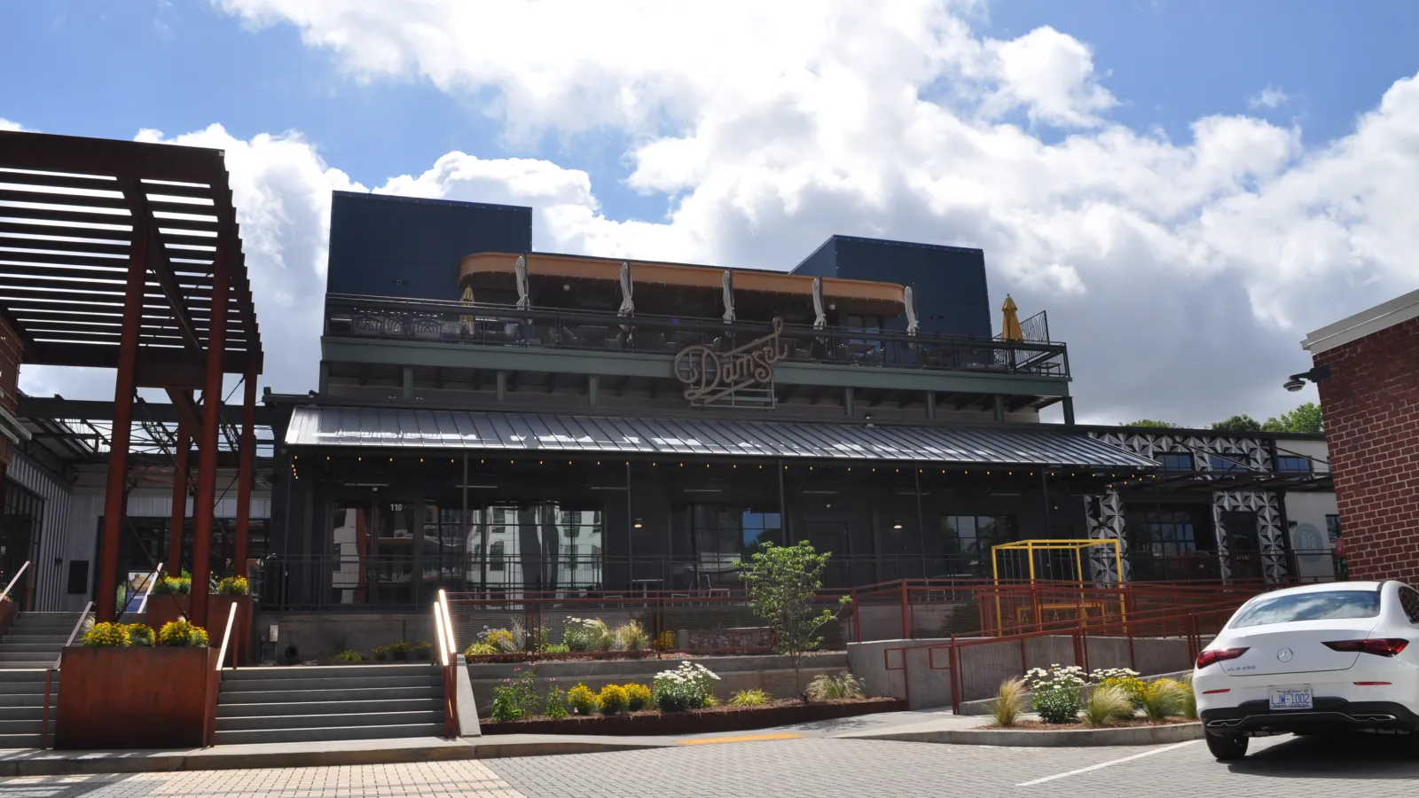 Modern two-story restaurant building with outdoor patios, greenery, and parked white car under a partly cloudy sky.