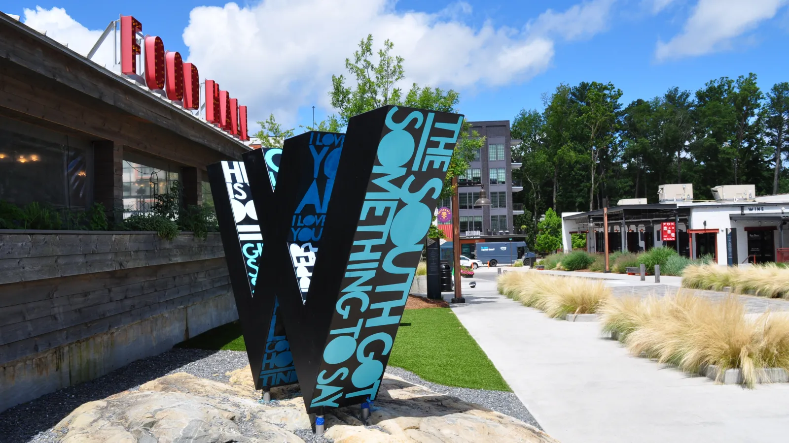 Large decorative 'W' sculpture with text in an outdoor urban park area under blue sky.