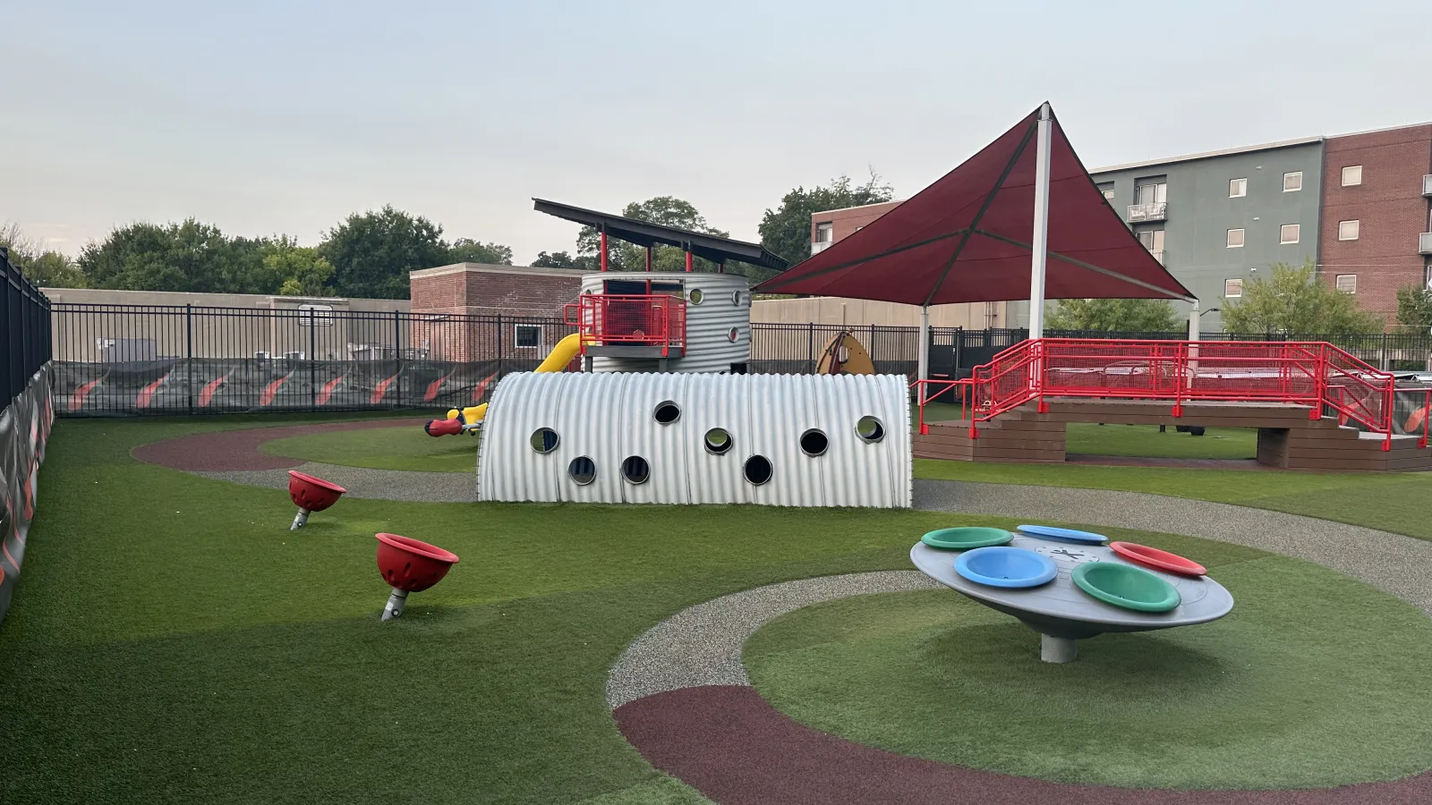 Modern outdoor playground with colorful spinning discs, slide, climbing structures, and shaded red canopy over seating area.