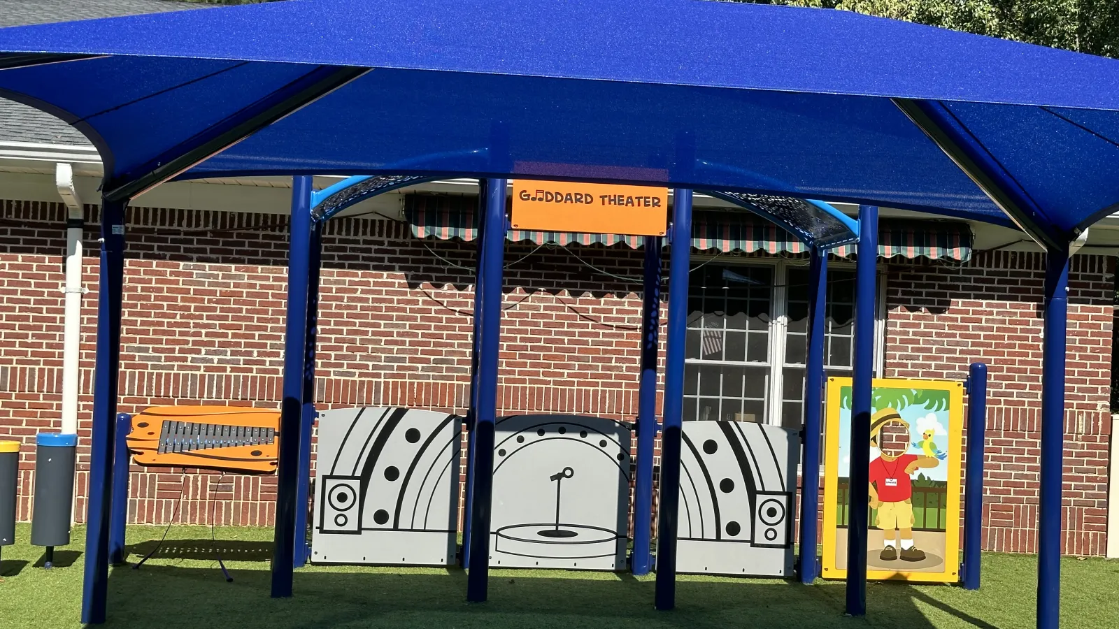 Outdoor children's play theater area with blue canopy, stage backdrop, and artificial grass seating resembling rocks.