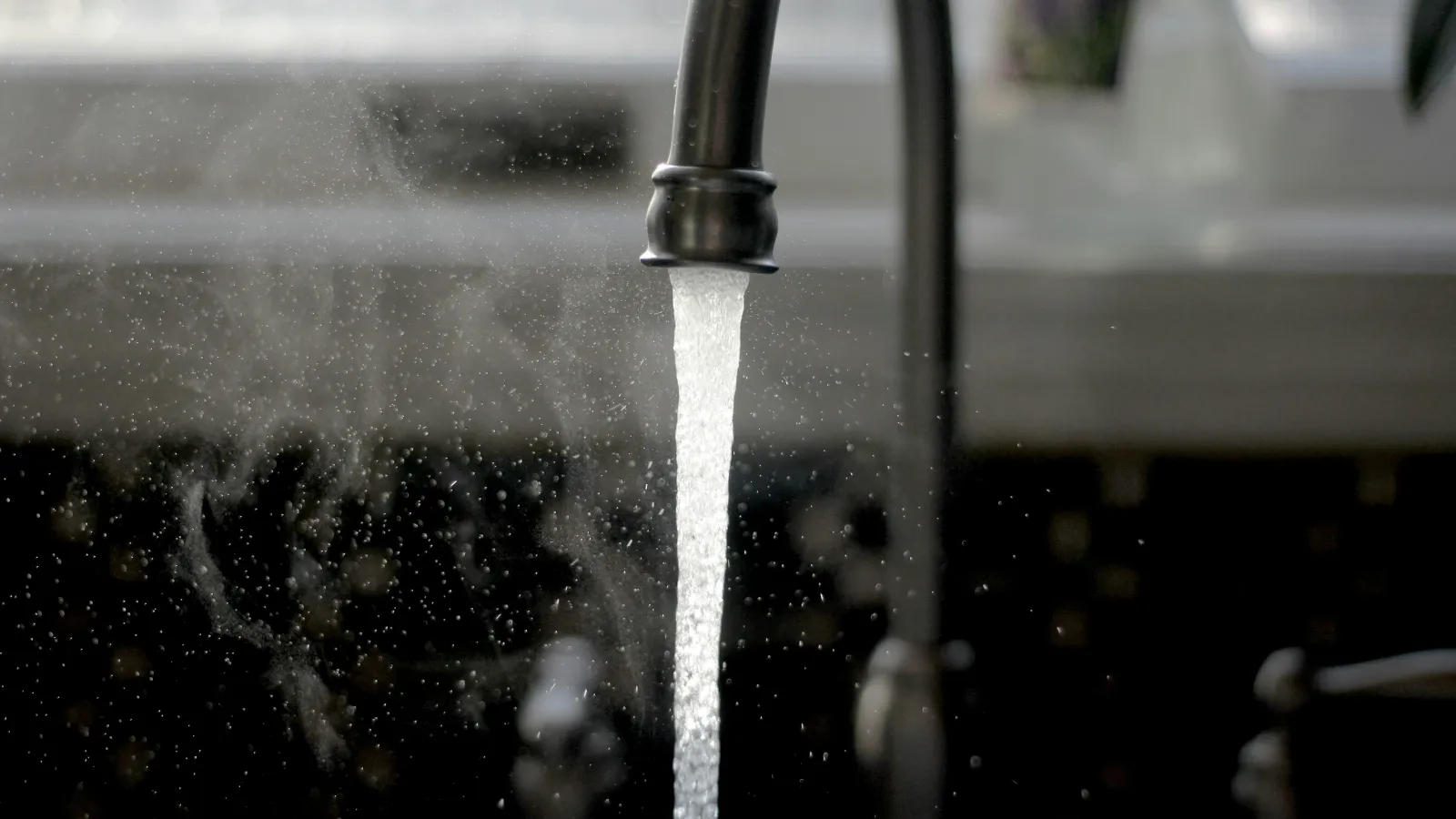 Steaming hot water flowing from a black kitchen faucet into a sink with blurred background.