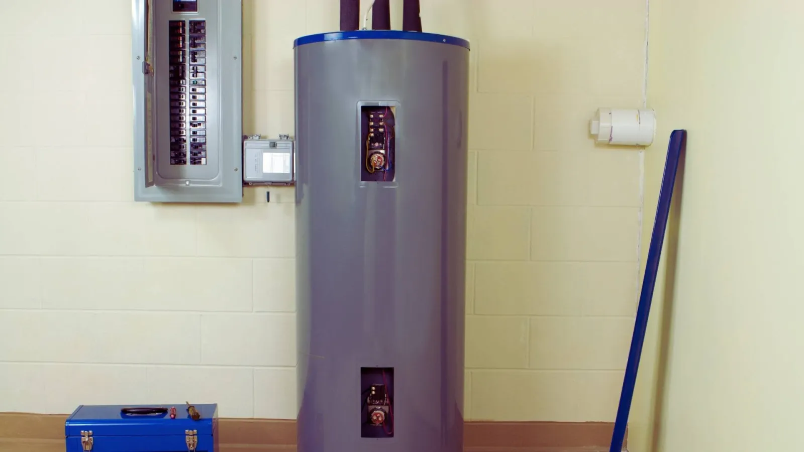 Gray water heater installed next to electrical panel inside a utility room with a blue toolbox nearby.