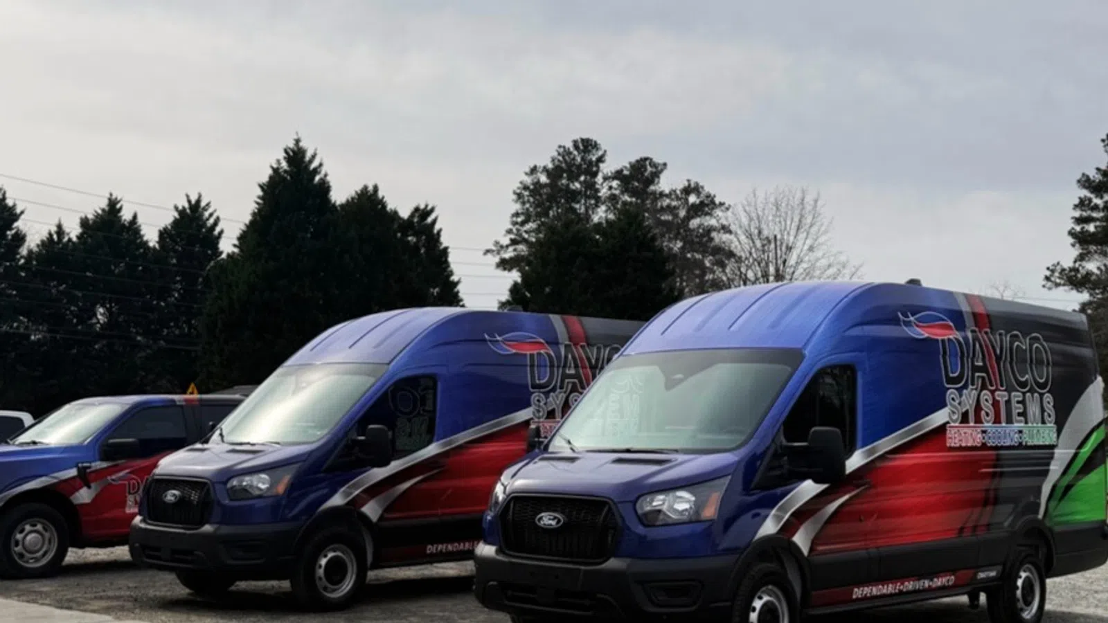 Two blue and red Ford vans with Dayco Systems branding parked outdoors on a cloudy day.