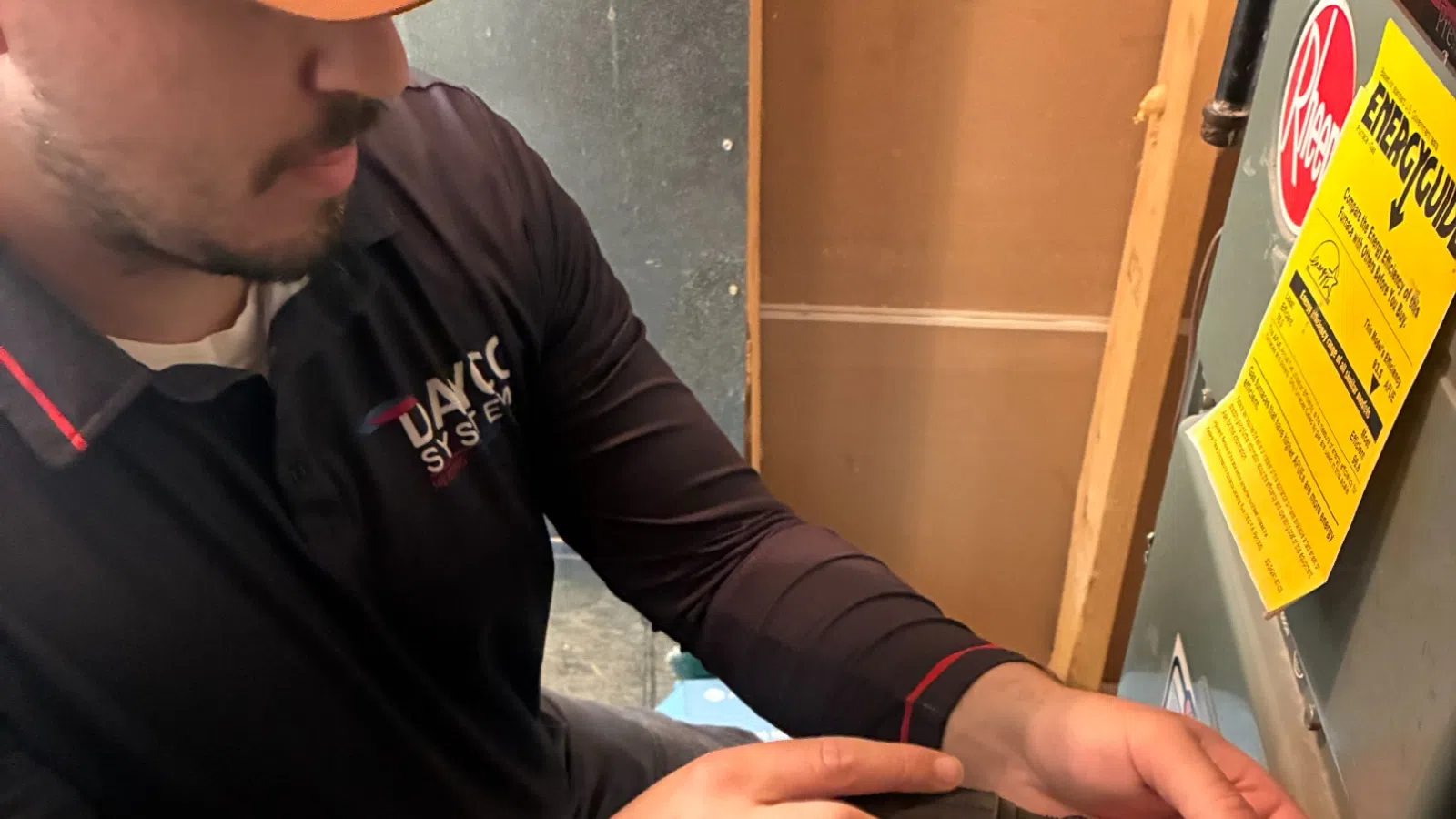 Technician wearing a brown cap and black shirt repairing an HVAC unit with a screwdriver indoors.