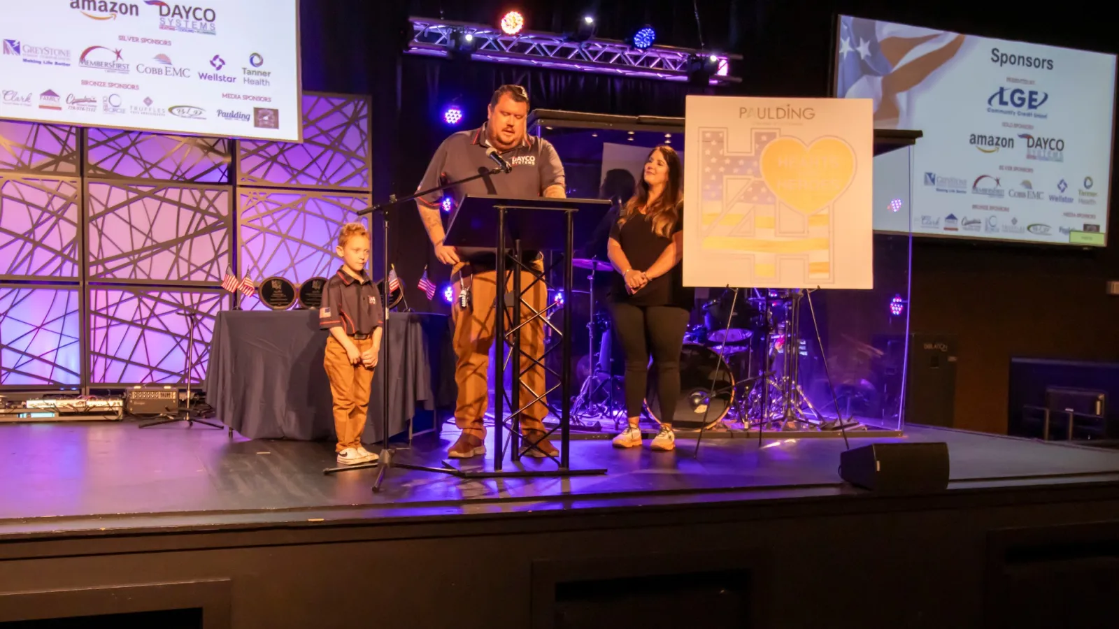 Man speaking at podium with woman and child standing beside him at event with sponsor logos and stage lighting.