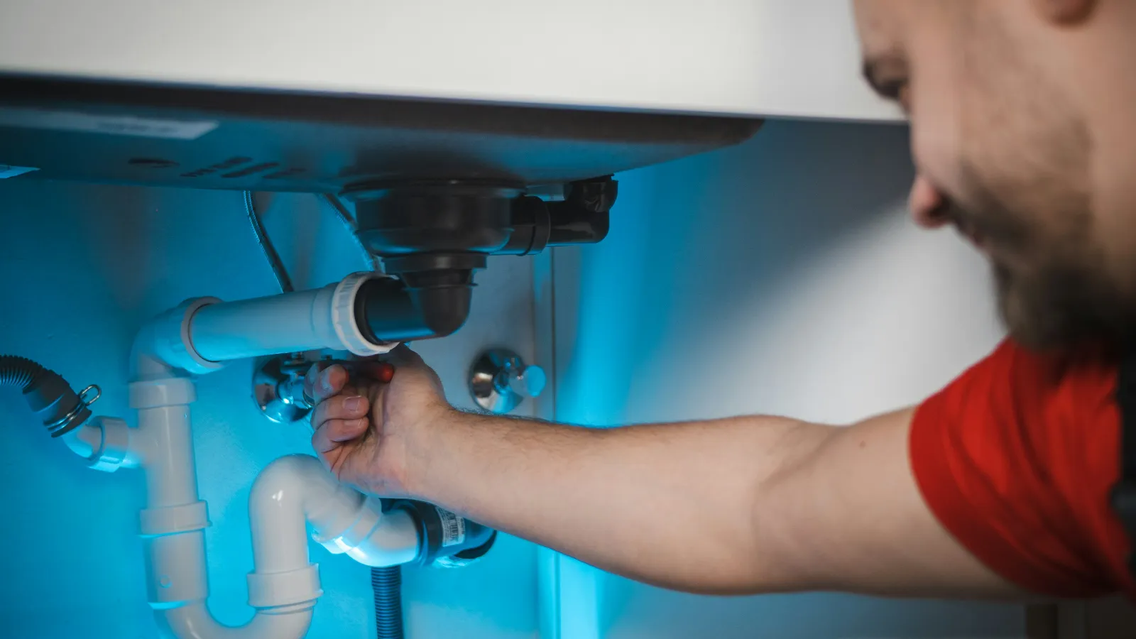 Man fixing white PVC plumbing pipes under a sink with a blue light illuminating the area.