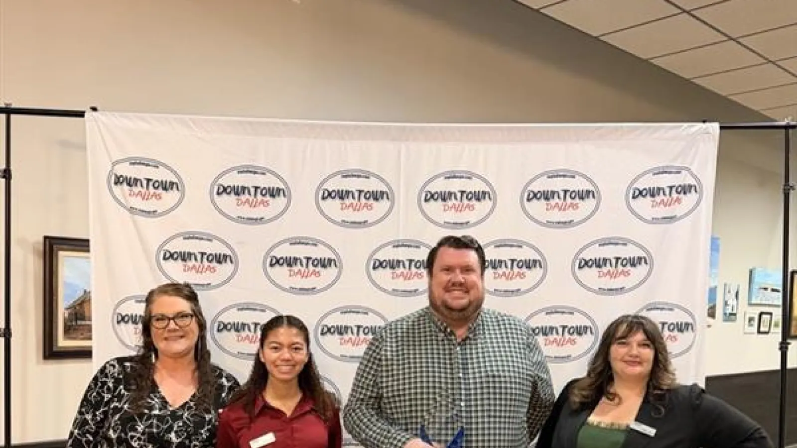 Four people standing in front of a Downtown Dallas backdrop at an indoor event, dressed in business casual attire.