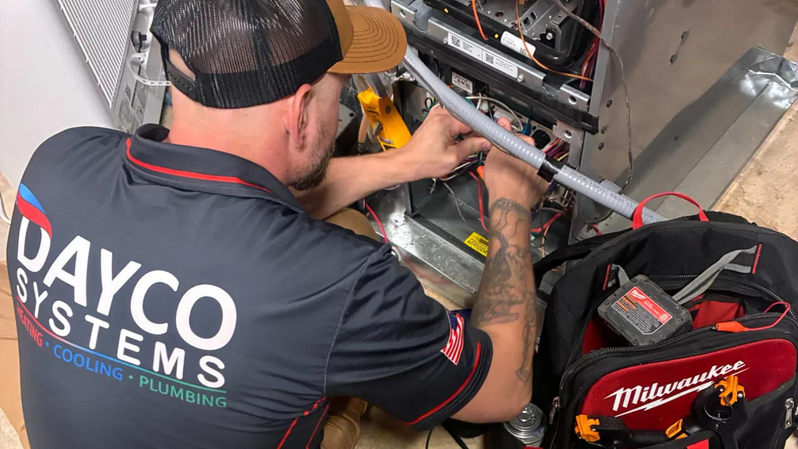 Technician in Dayco Systems shirt repairing HVAC unit with Milwaukee tool bag nearby on a concrete floor.