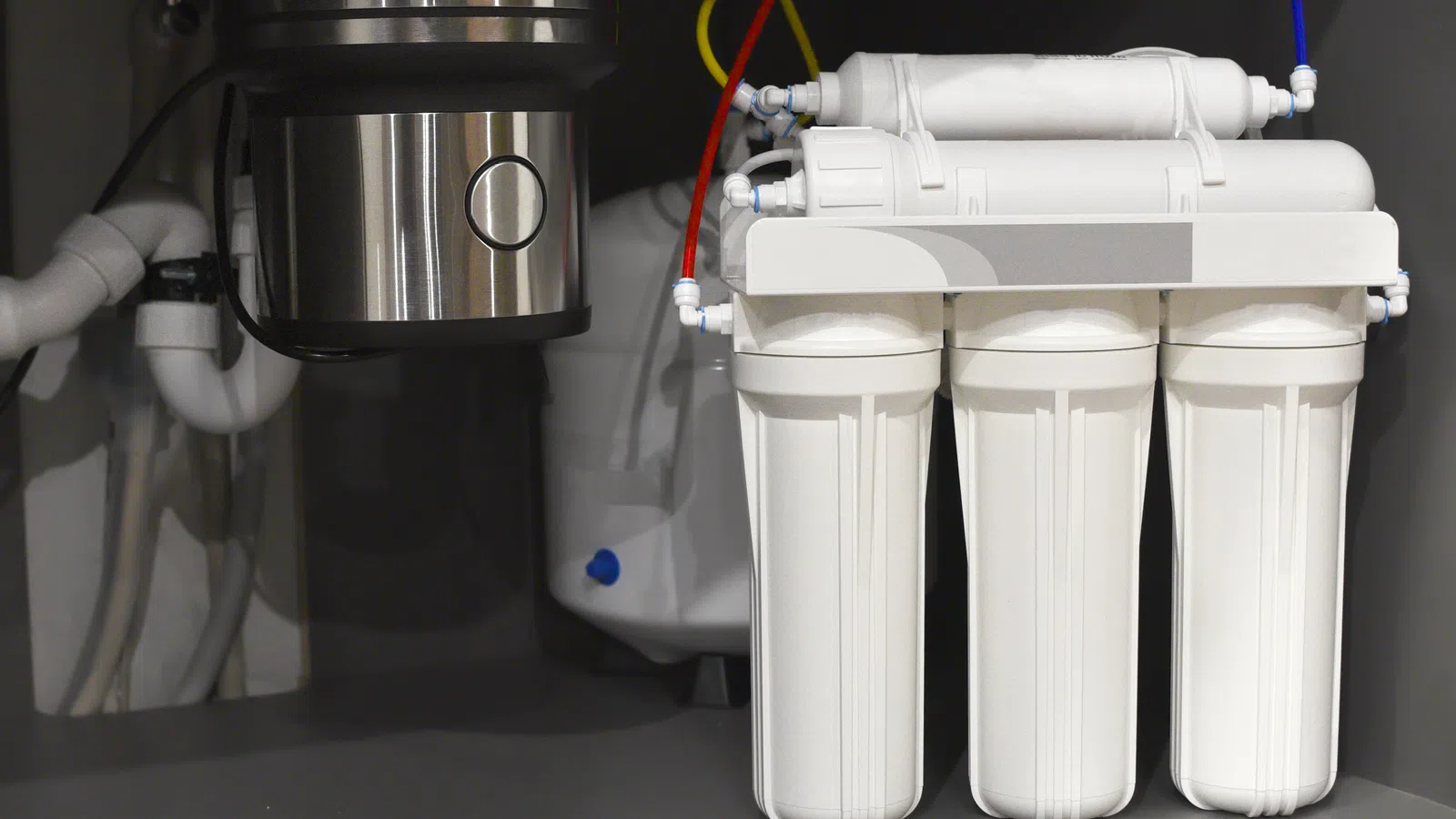 Under-sink reverse osmosis water filtration system with white canisters and colored tubing installed beside plumbing.