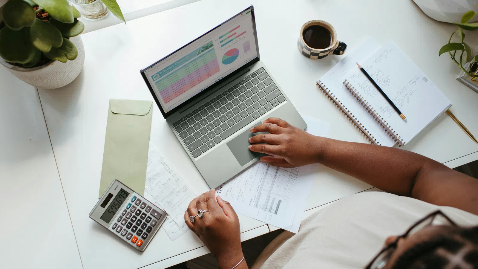 Person using a laptop to analyze financial data, with calculator, paperwork, notebook, and coffee on desk.