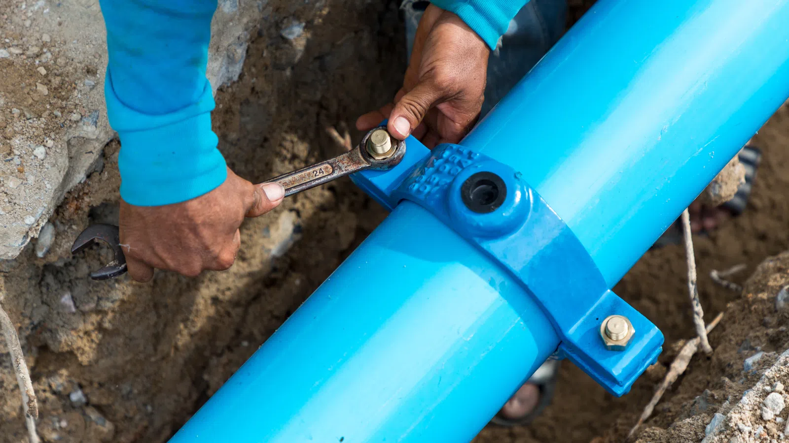 Worker tightening bolt on large blue underground water pipe using wrench in construction site trench