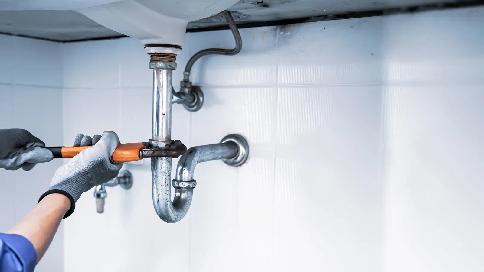 Plumber wearing gloves fixing a pipe under a sink with a wrench in a tiled bathroom setting.