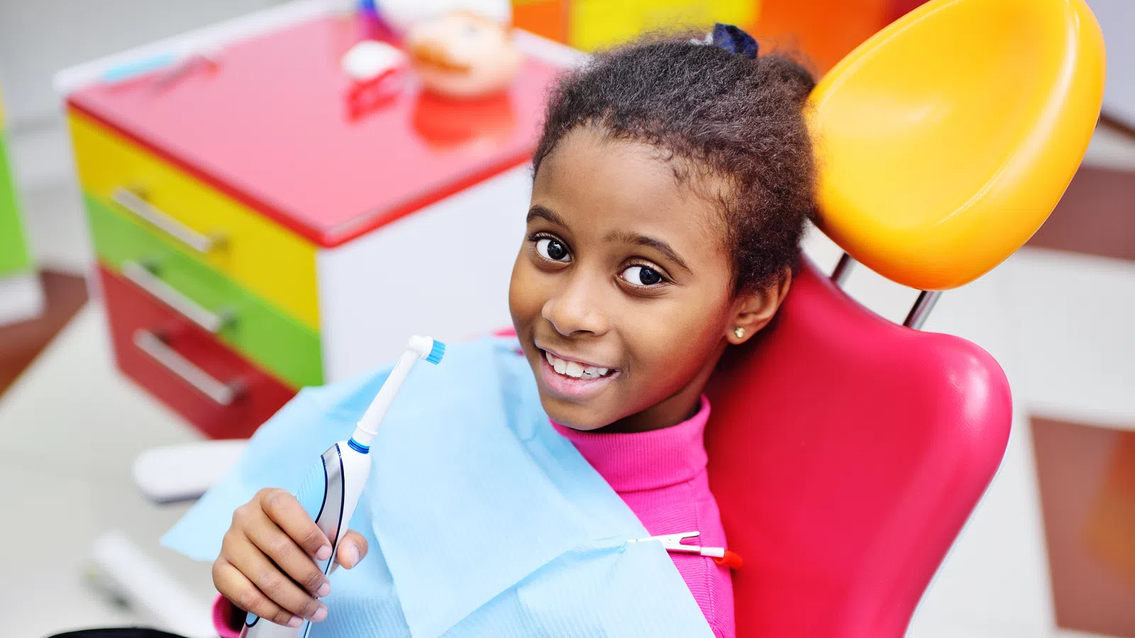 Smiling young girl sitting in a colorful dental chair holding an electric toothbrush at the dentist office.