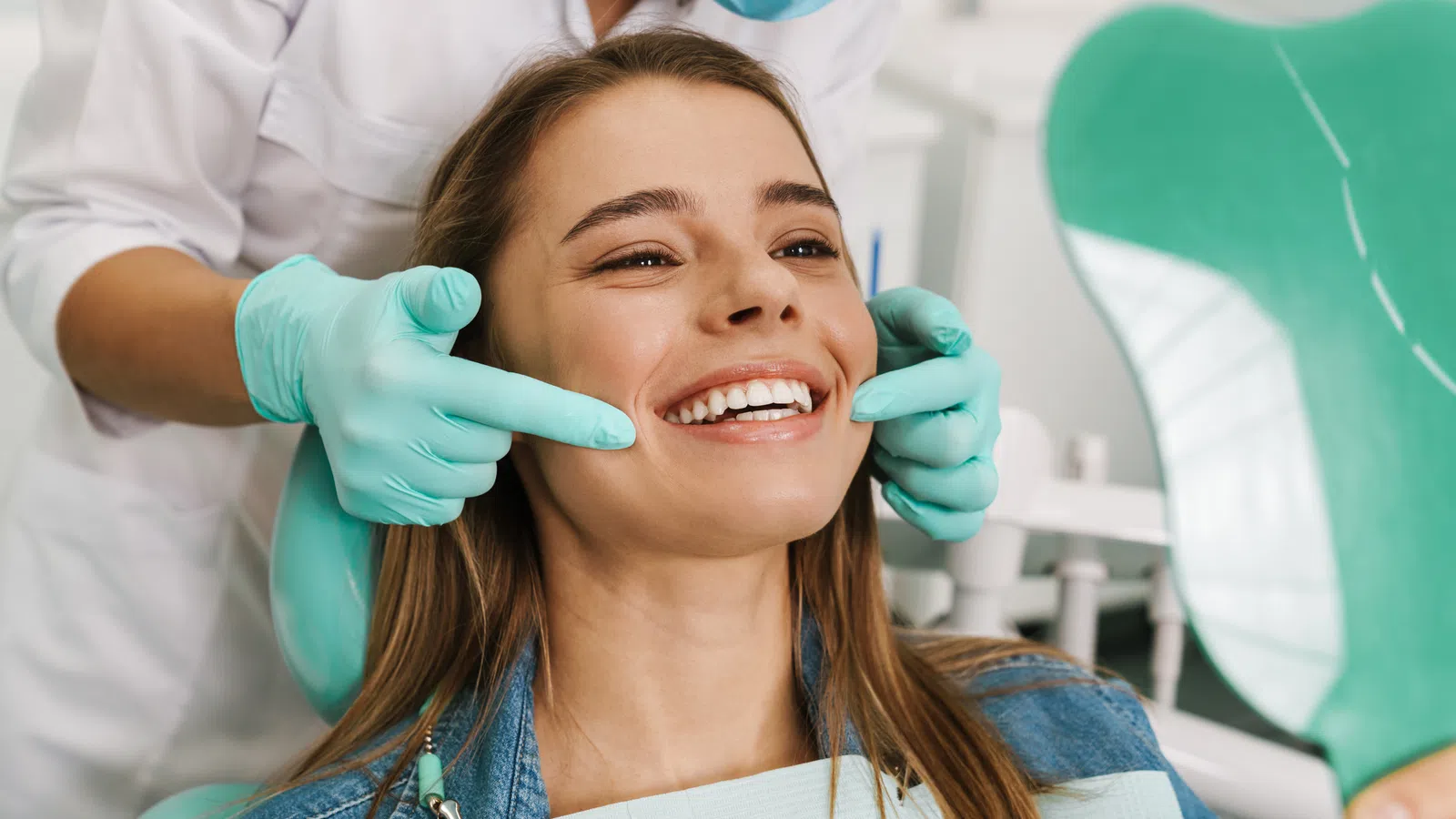 Dentist wearing gloves examines a smiling woman's teeth using a mirror in a dental clinic.