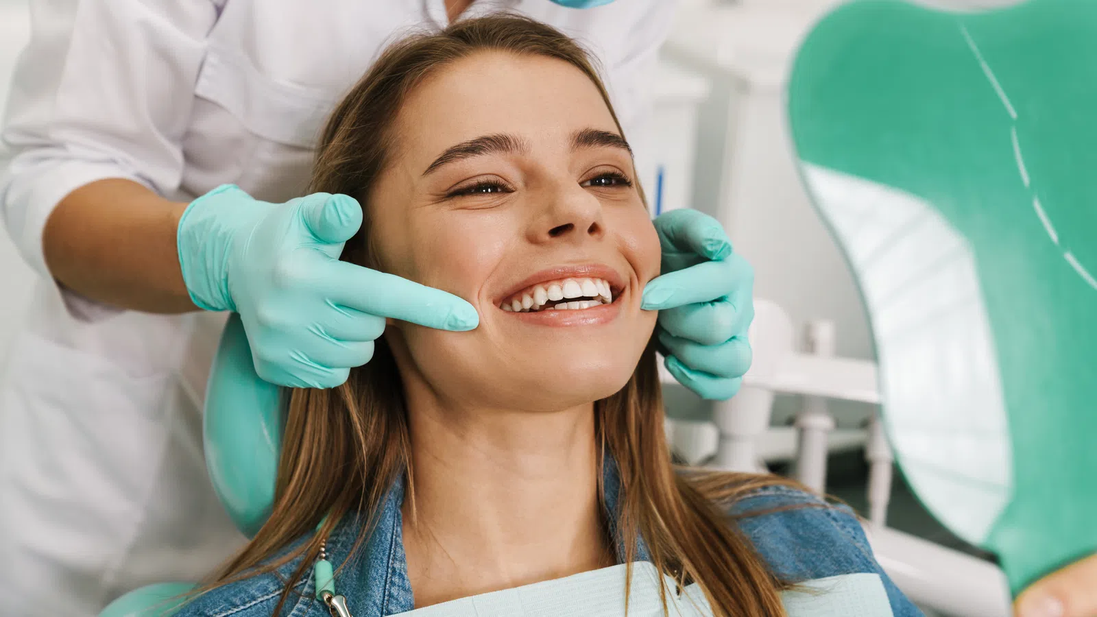 Dentist wearing gloves examines a smiling woman's teeth using a mirror in a dental clinic.