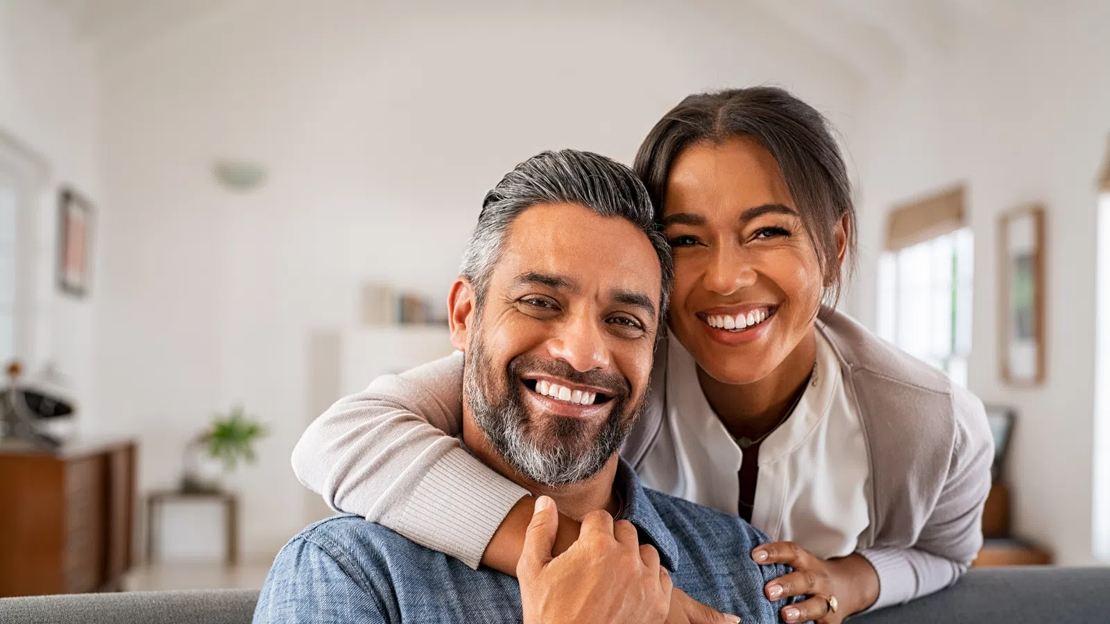 Happy couple smiling and hugging on a couch in a bright living room with cozy decor and natural light.