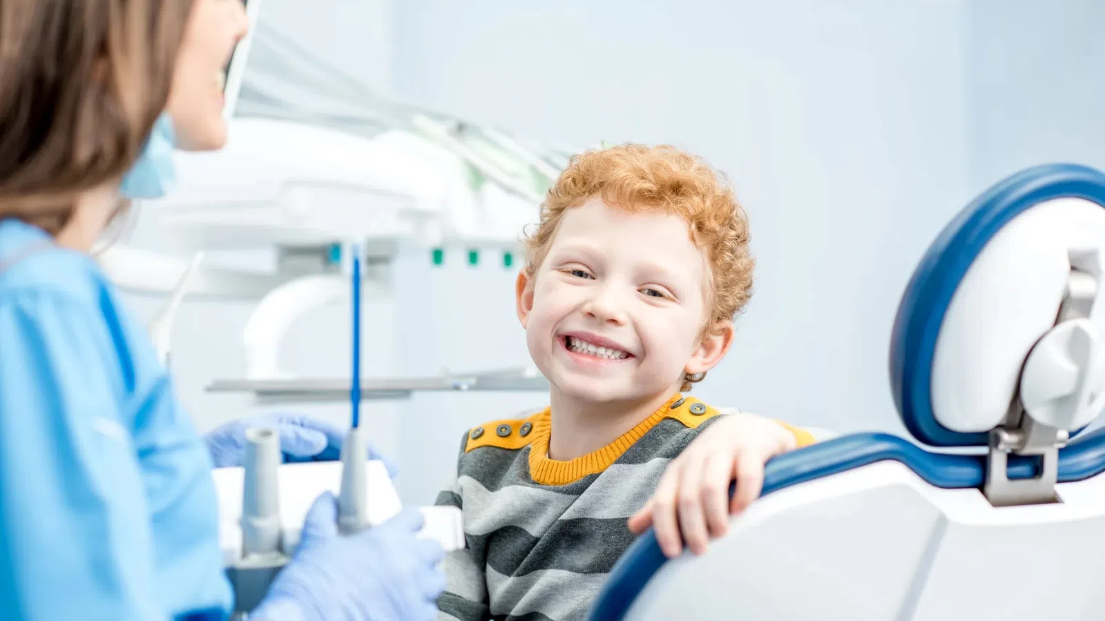 Smiling red-haired boy sitting in dental chair with dentist in clinic, dental equipment visible in background