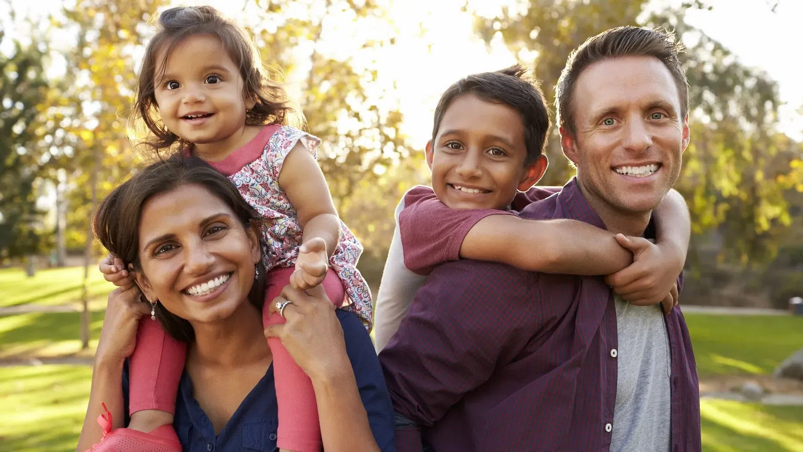 Smiling diverse family outdoors with parents carrying their children on shoulders in sunlit park setting.