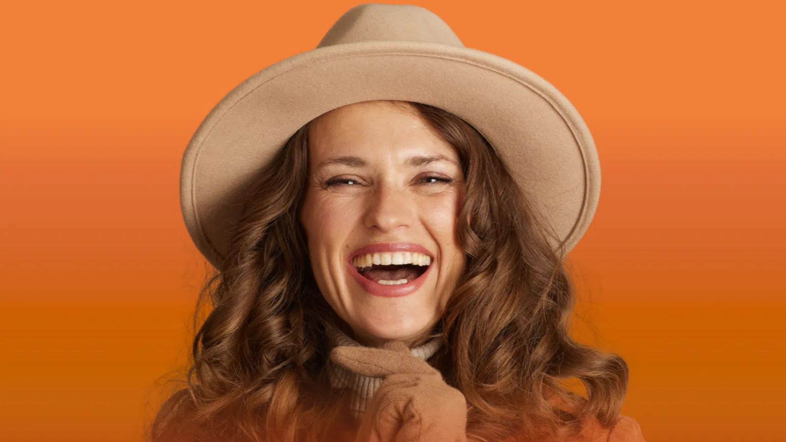 Smiling woman with curly hair wearing beige hat and gloves in front of an orange gradient background