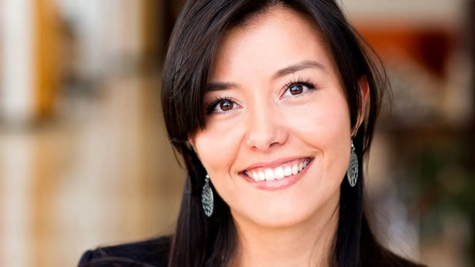 Smiling young woman with dark hair and earrings in a softly lit indoor setting with blurred background.