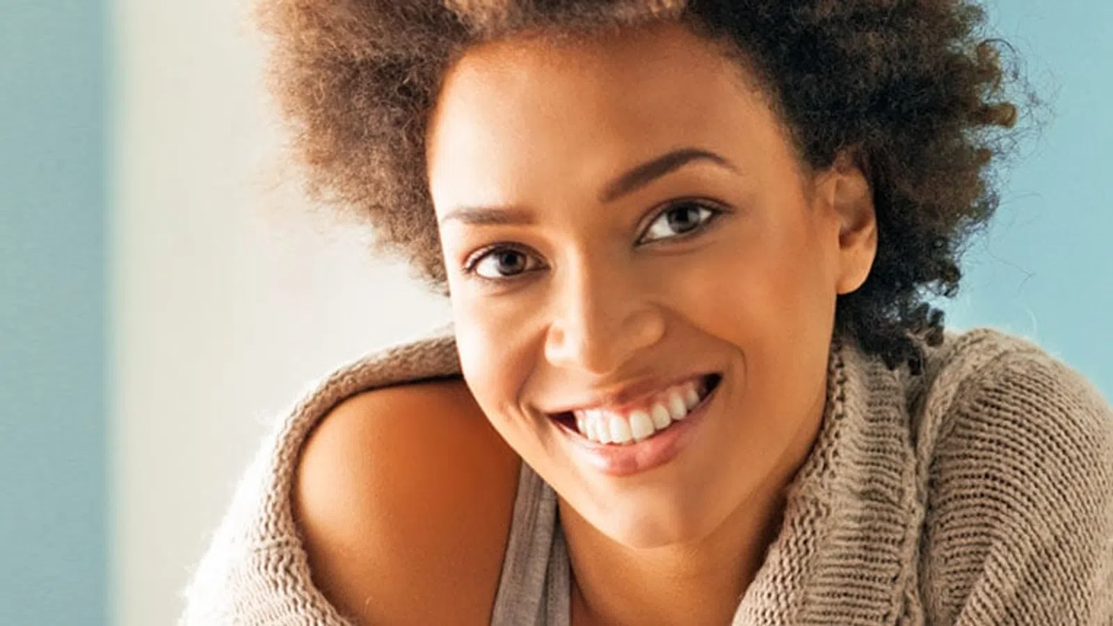 Smiling woman with curly hair wearing a cozy beige sweater against a light blue background