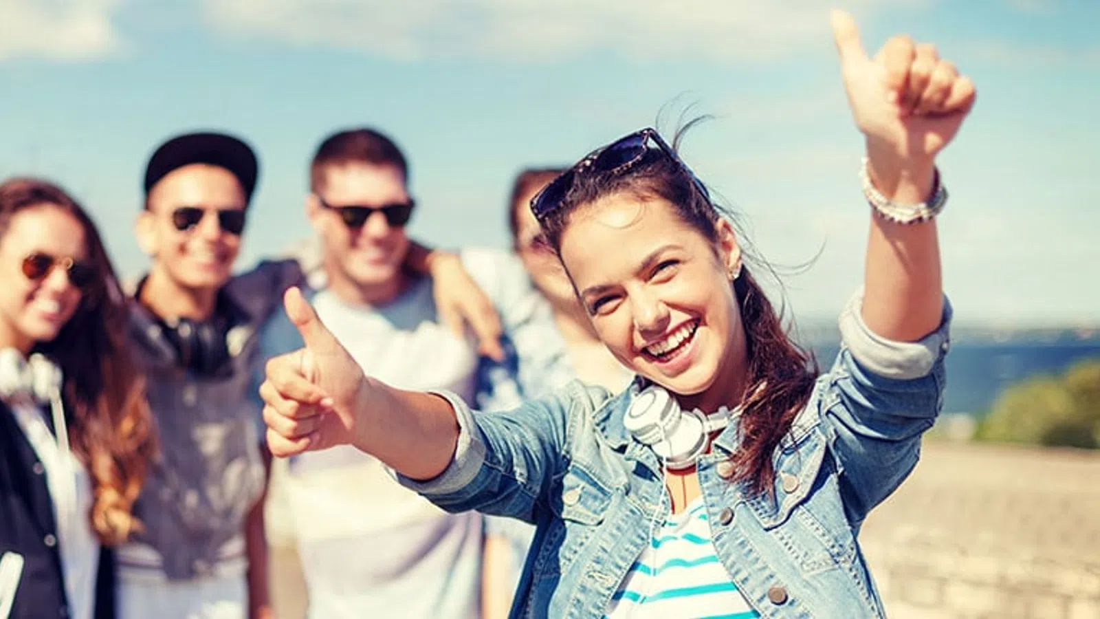 Group of young friends smiling outdoors with a sunny sky, girl in front giving thumbs up and wearing headphones.