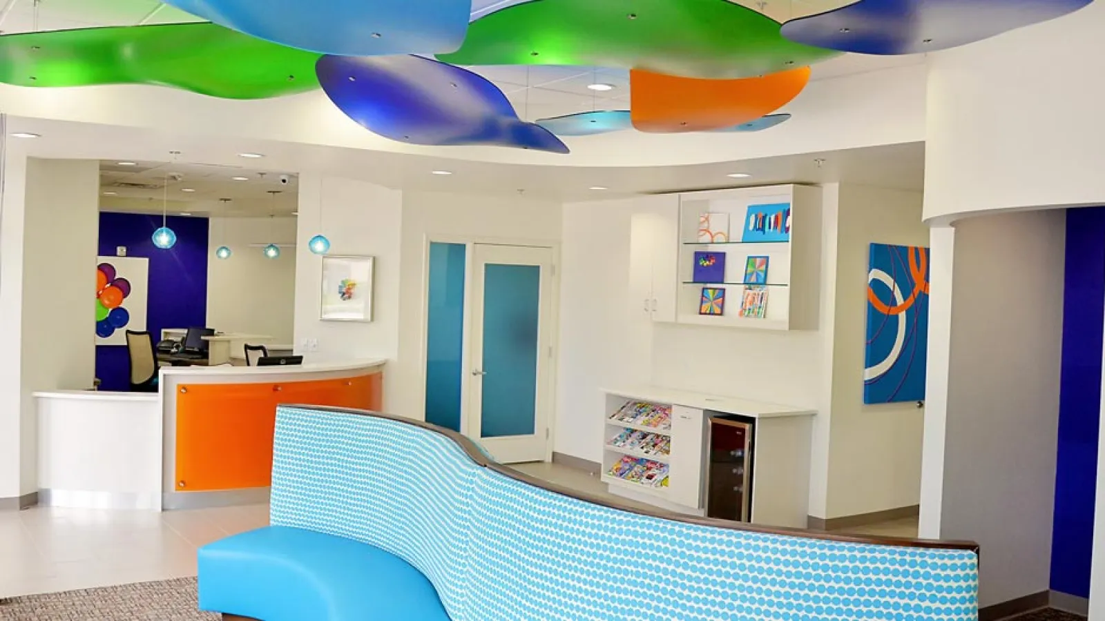 Bright modern waiting room with colorful ceiling panels, curved blue seating and orange-accented reception desk.