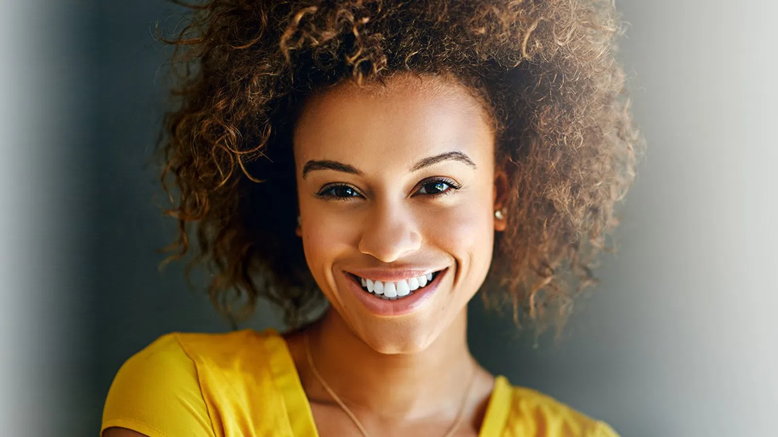 Smiling woman with curly hair wearing a yellow top against a blurred gray background.