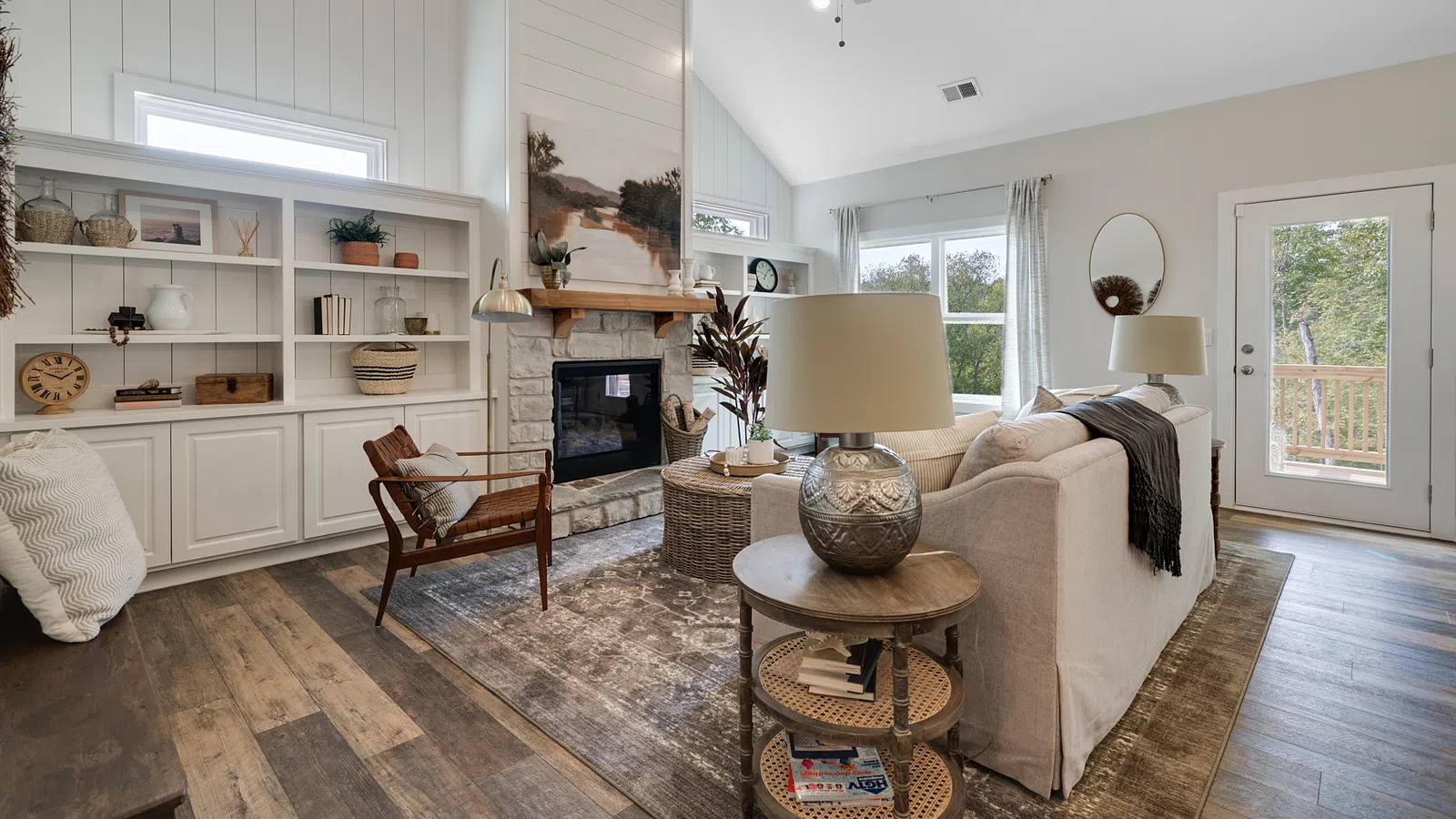 Bright living room with vaulted ceiling, stone fireplace, wooden furniture, and large windows overlooking greenery.