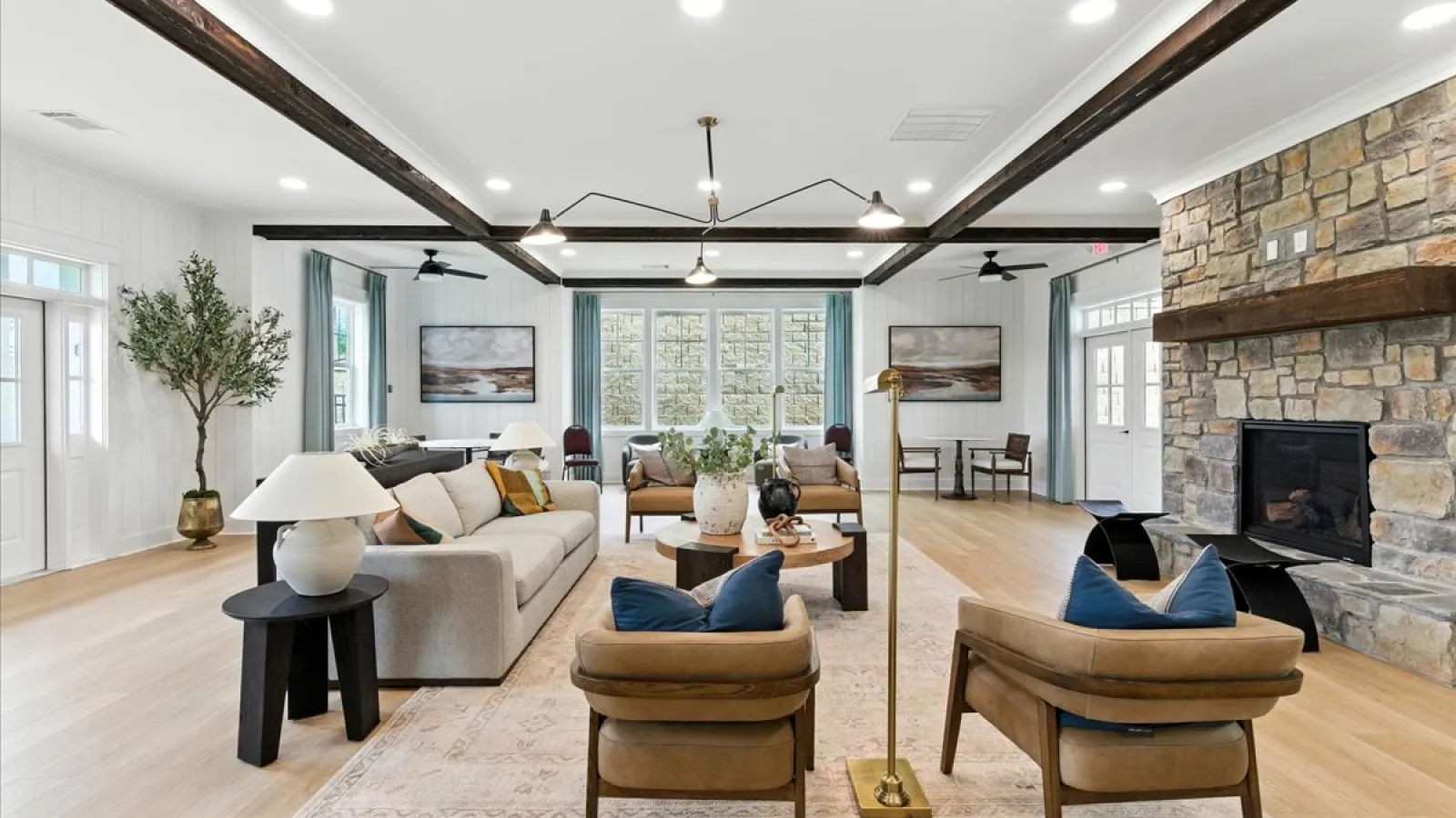 Spacious living room with beige sofas, brown chairs, stone fireplace, wooden beams, and large windows letting in natural light