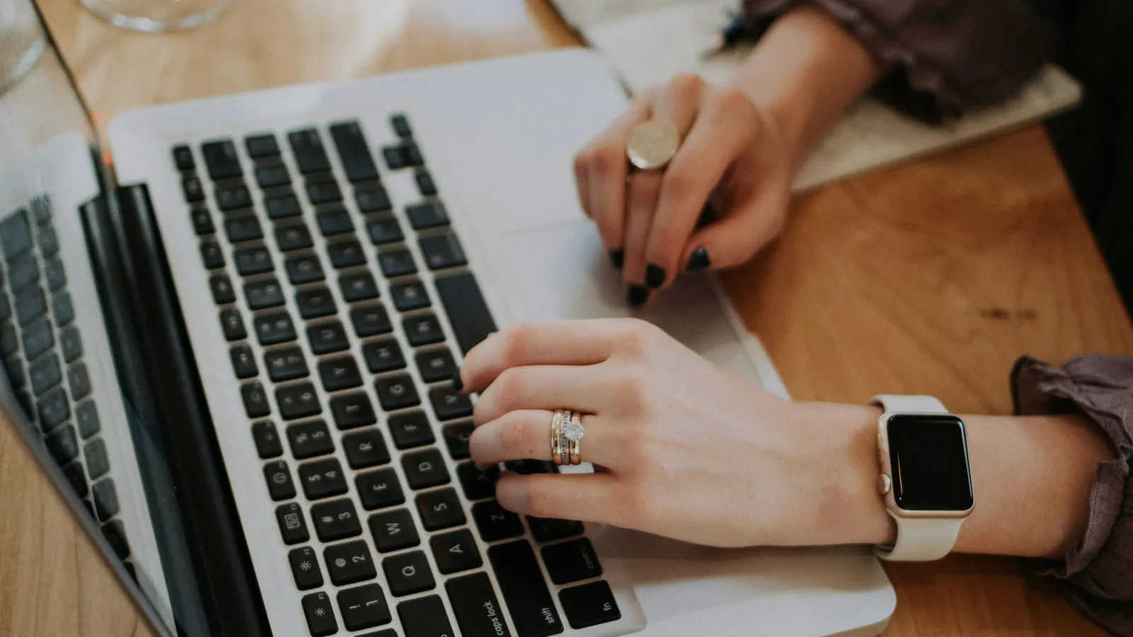 Person wearing rings and a smartwatch typing on a laptop keyboard over a wooden desk.