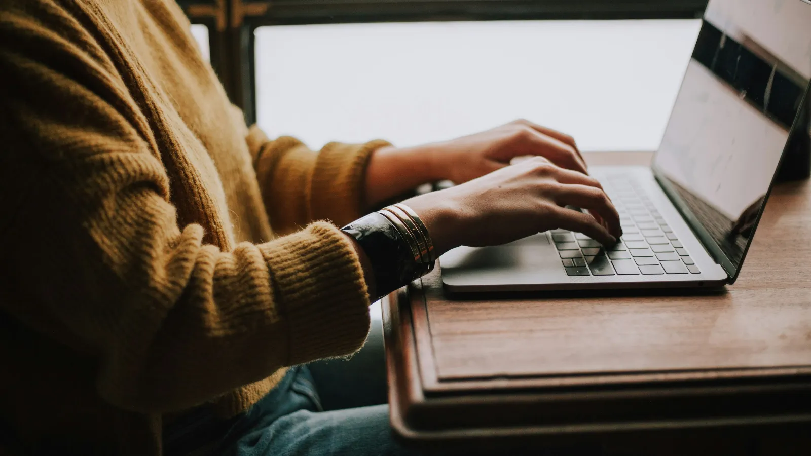 Person in a brown sweater typing on a laptop at a wooden desk near a window with natural light.