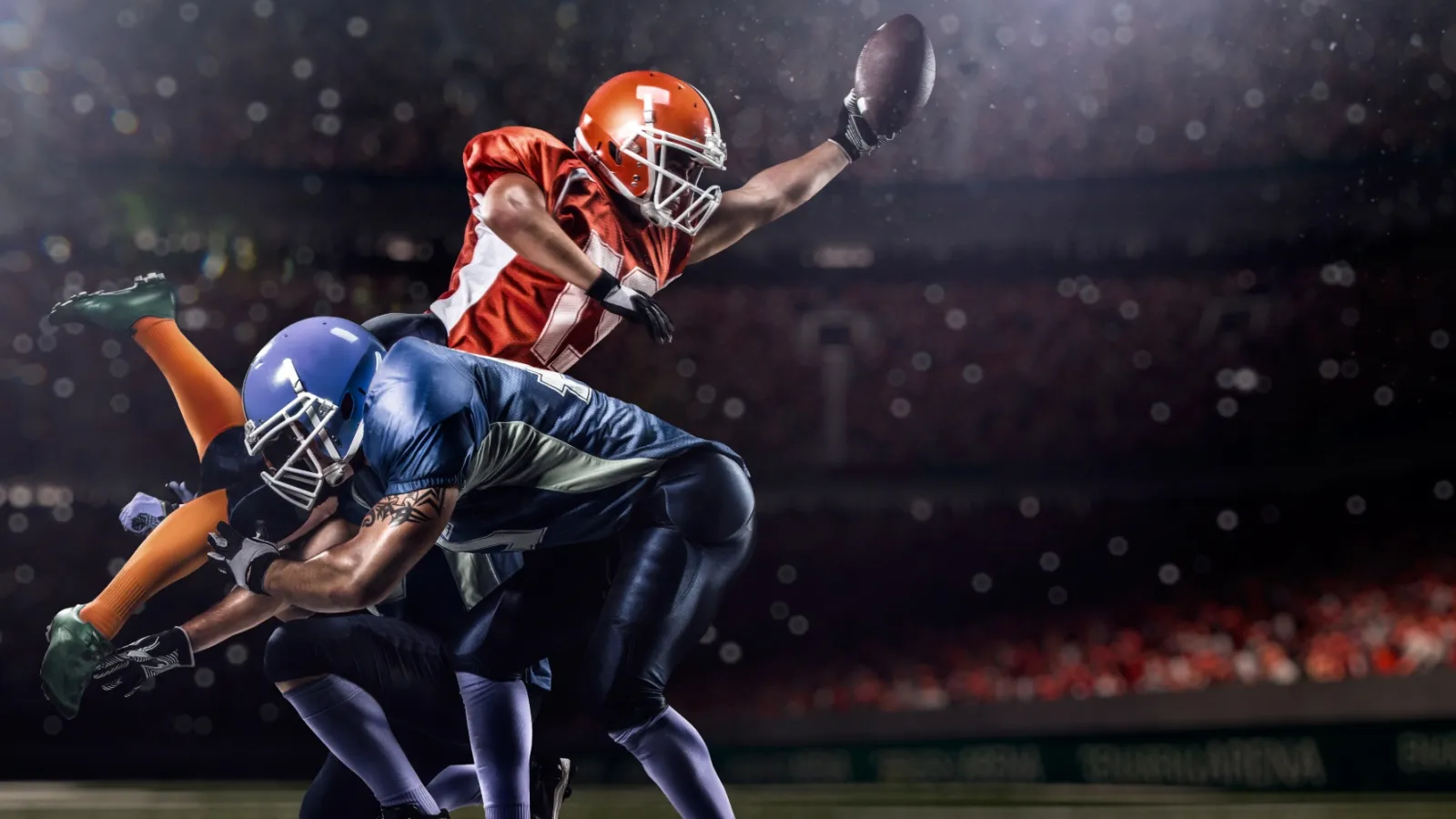 Football player in orange jersey reaches to catch ball while blue jersey defender tackles him on nighttime field.
