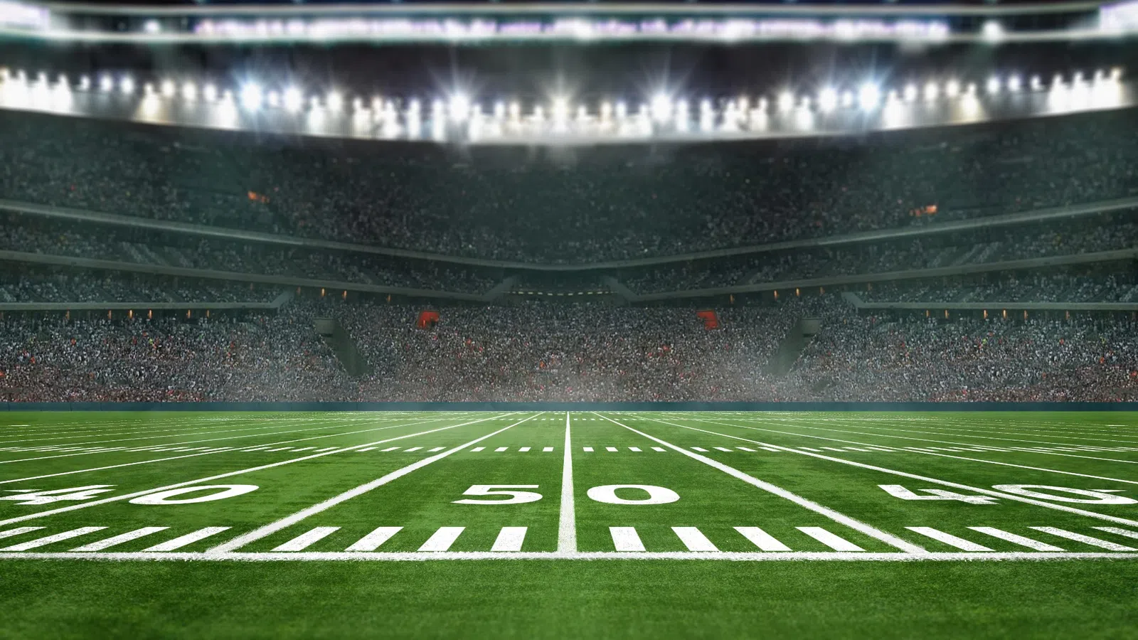 View of a brightly lit American football stadium with a large crowd and marked green field at the 50-yard line.