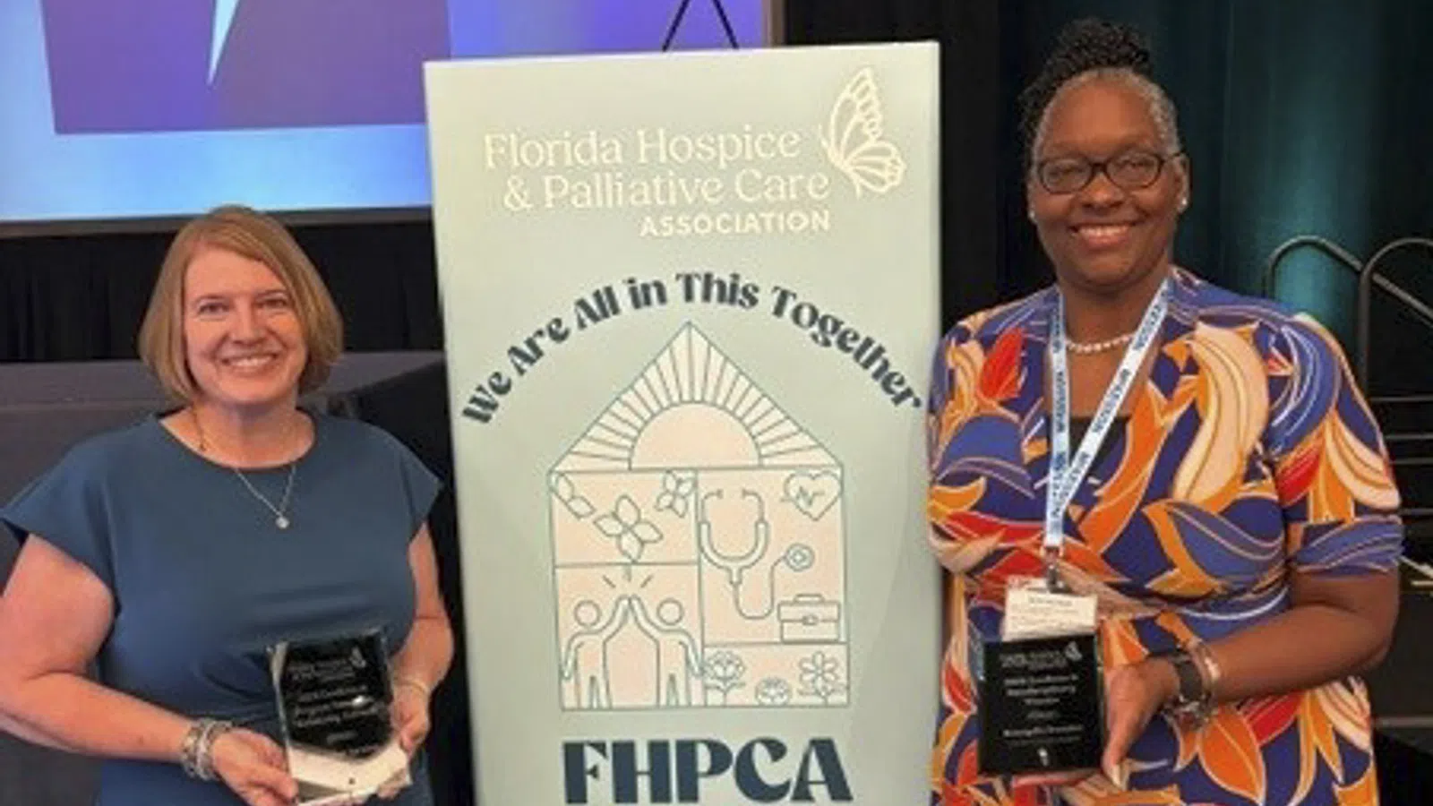 Two women holding awards next to a sign for the Florida Hospice & Palliative Care Association Annual Forum