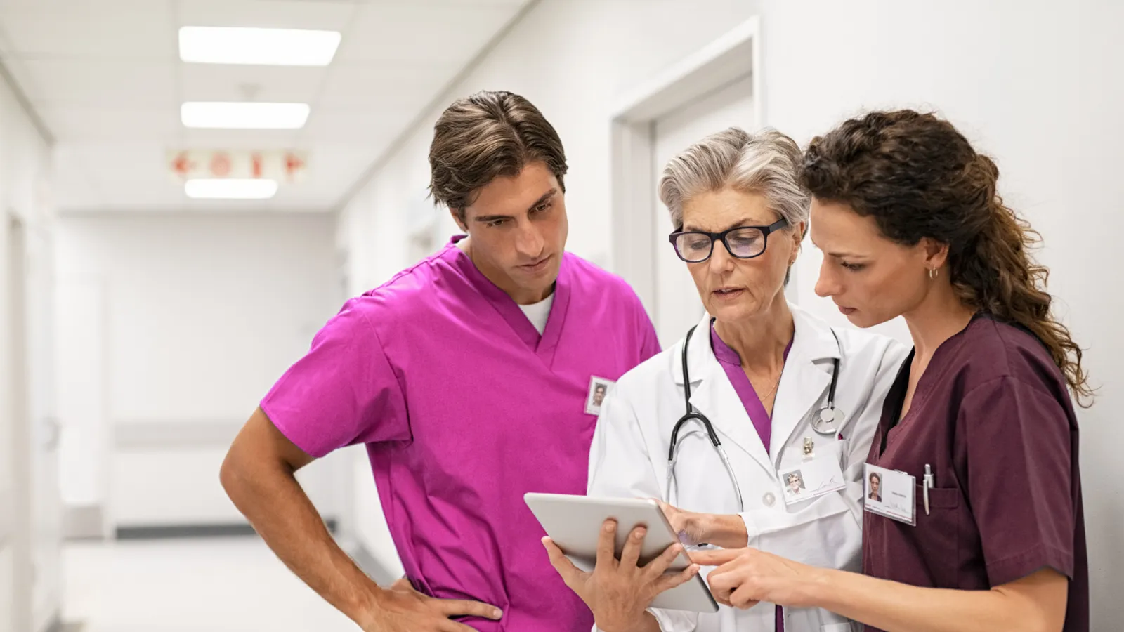 Three healthcare professionals discussing patient information on a digital tablet in a hospital hallway.