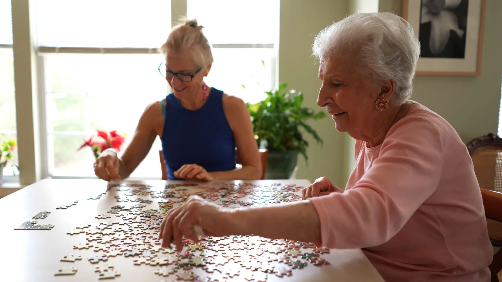 Two elderly women happily assembling a large jigsaw puzzle together at a sunlit table indoors