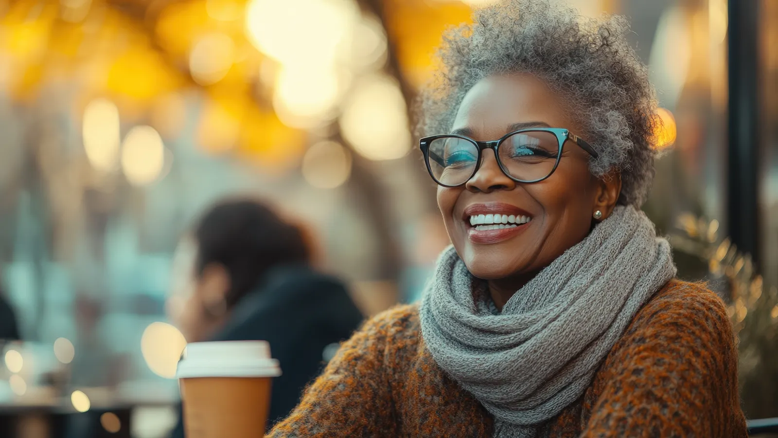Smiling older woman with gray hair and glasses wearing a scarf and sweater at a cafe in autumn.