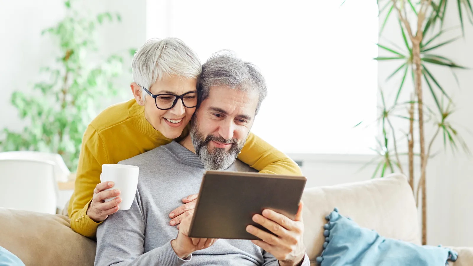 Senior couple enjoying time together on a couch using a tablet, woman holding a coffee mug and smiling.