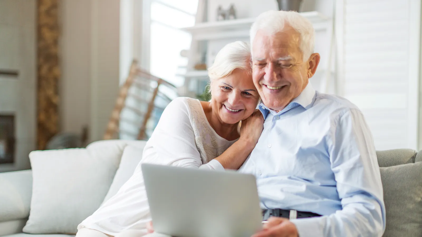 Smiling elderly couple sitting on a couch and using a laptop together in a bright living room.